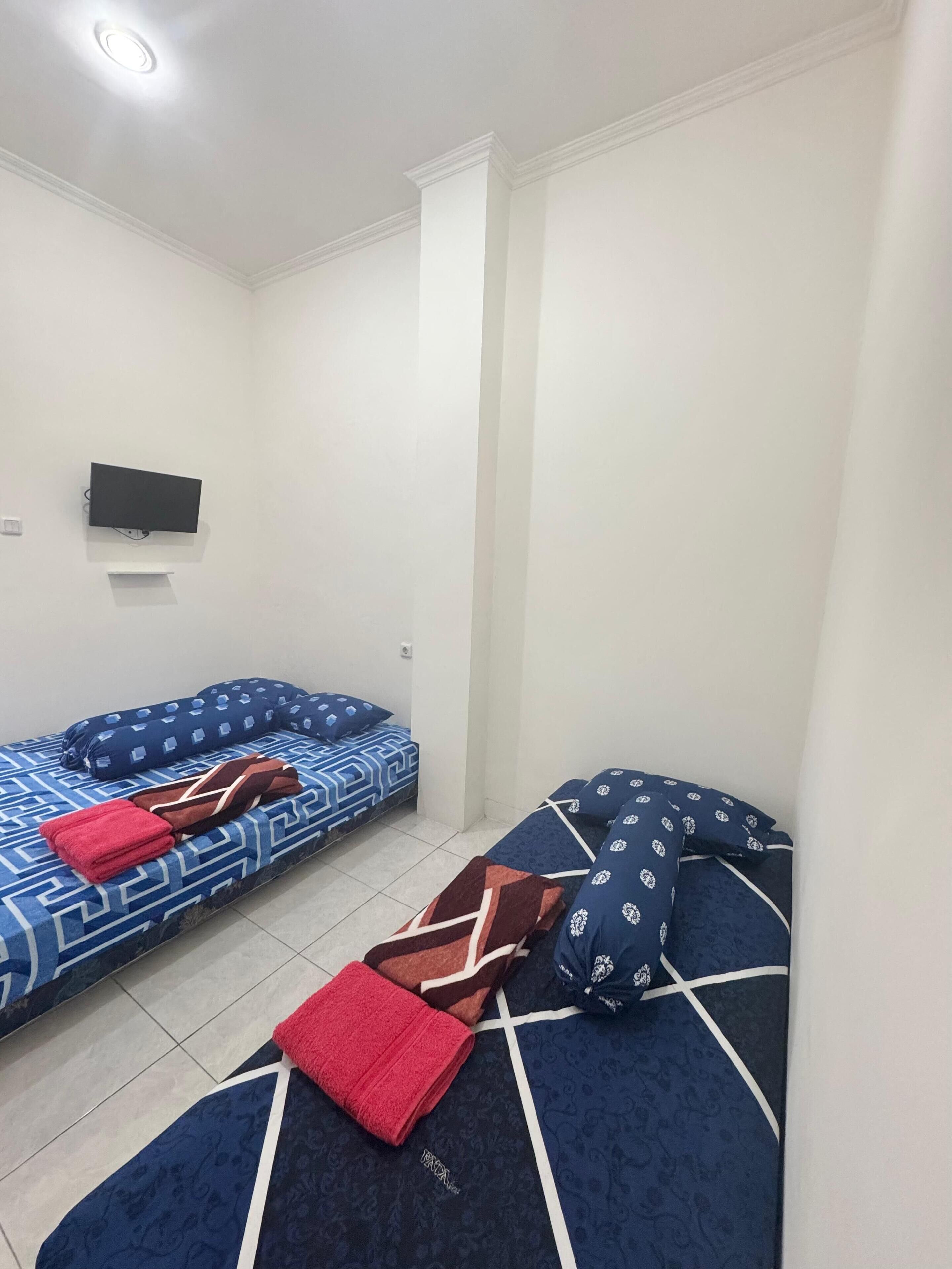 Ein kleiner, sauberer Raum mit zwei Betten, jeweils mit gemusterten Bettbezügen und einem roten Handtuch. Ein Flachbildfernseher ist an der Wand montiert.