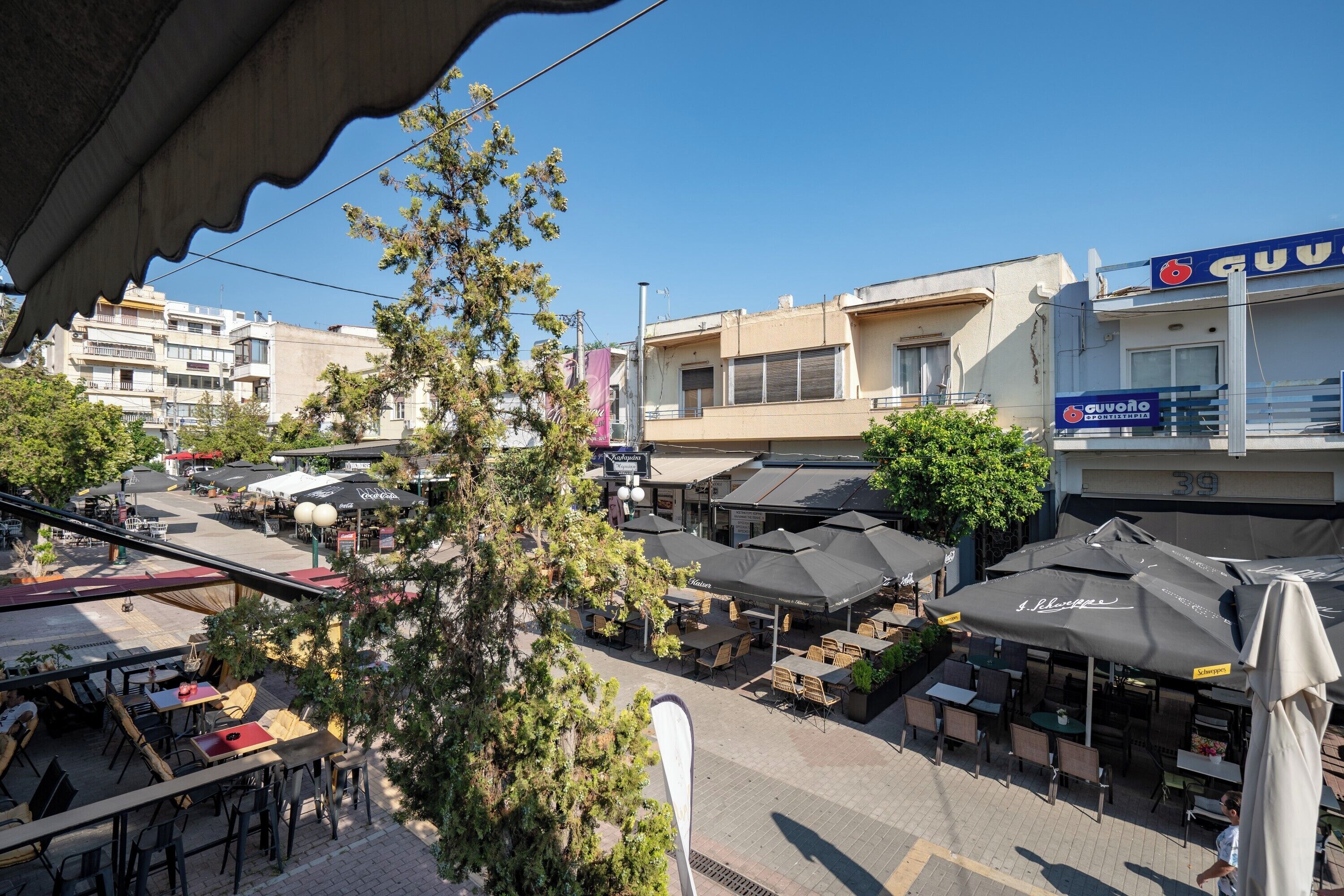 Café al aire libre con mesas y sombrillas, rodeado de edificios y árboles.