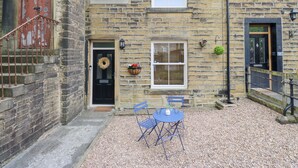 A stone building with a black door, a small garden with a blue table and chairs, and a gravel path. - Hivewood Cottage (Keighley)