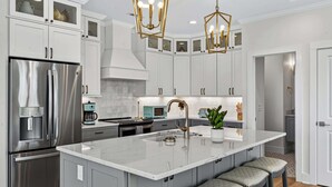 A modern kitchen with a large island, stainless steel appliances, and a unique geometric chandelier. - The Pointe of No Return (Ocean Isle Beach)
