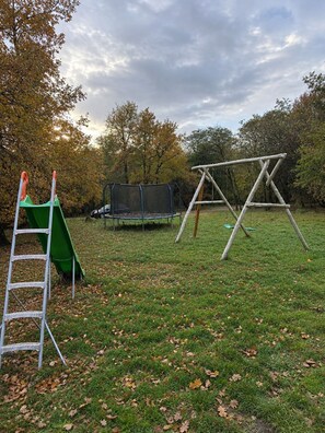 Un espacio al aire libre con césped, un parque infantil con columpio, tobogán y cama elástica.