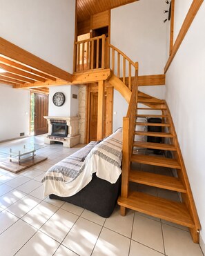 Amplia habitación con escalera de madera que conduce a un nivel superior, chimenea y cama con manta.