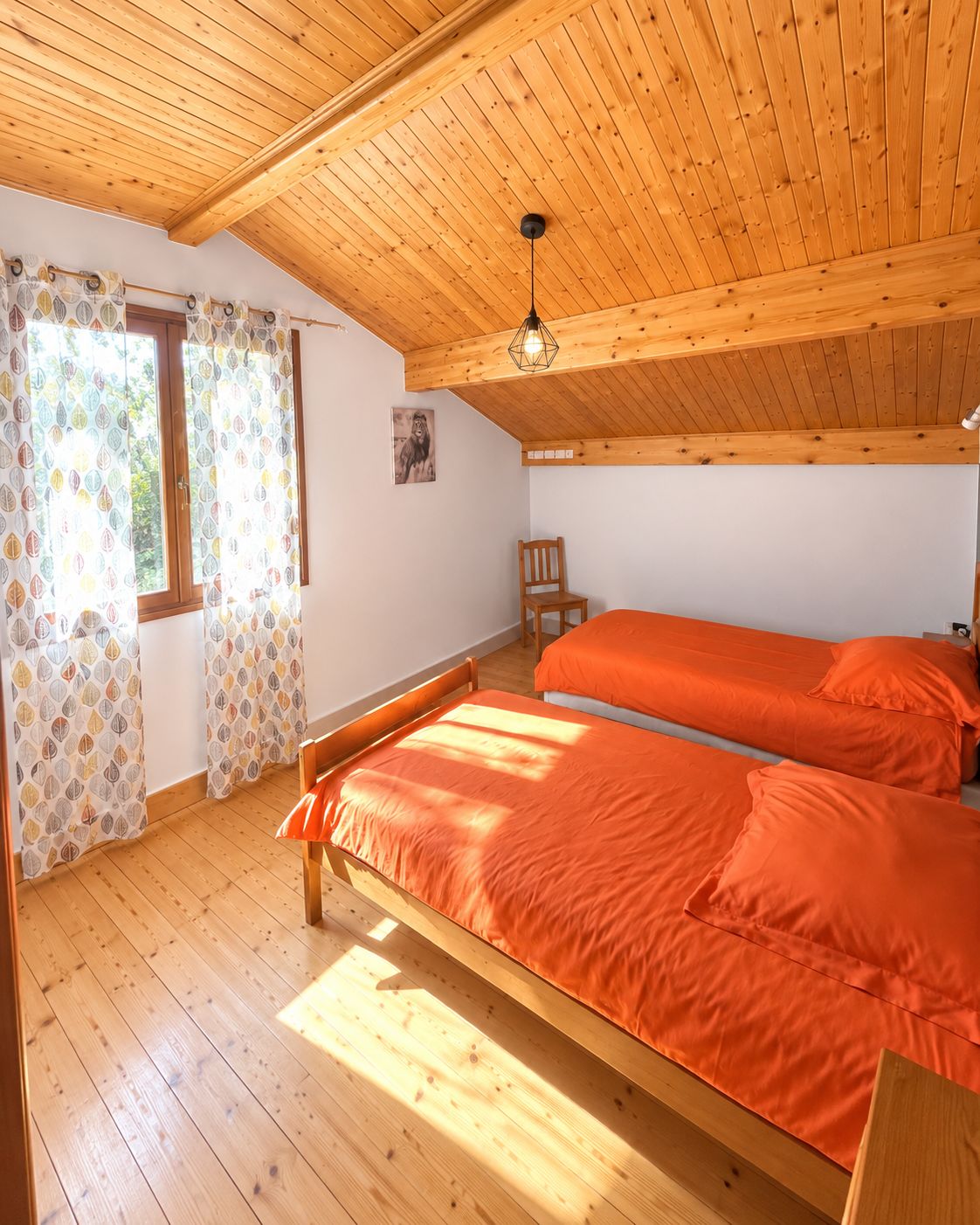 Une pièce avec un plancher en bois, un lit avec une literie orange, une chaise et une fenêtre ornée de rideaux à motifs.
