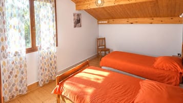 Une pièce avec un plancher en bois, un lit avec une literie orange, une chaise et une fenêtre ornée de rideaux à motifs.