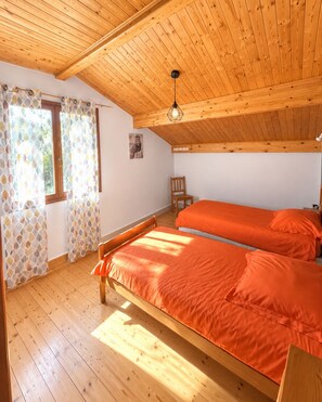 A room with wooden flooring, a bed with orange bedding, a chair, and a window with patterned curtains.
