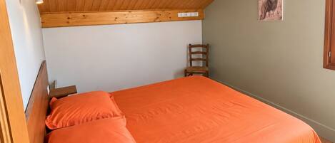 Une chambre avec un plafond en bois, un lit recouvert d’une couverture orange, une chaise en bois et un luminaire fixé au mur.