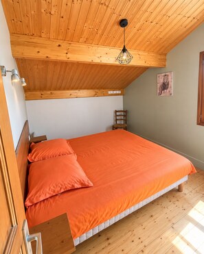 Un dormitorio con techo de madera, una cama con colcha naranja, una silla de madera y una lámpara fijada a la pared.