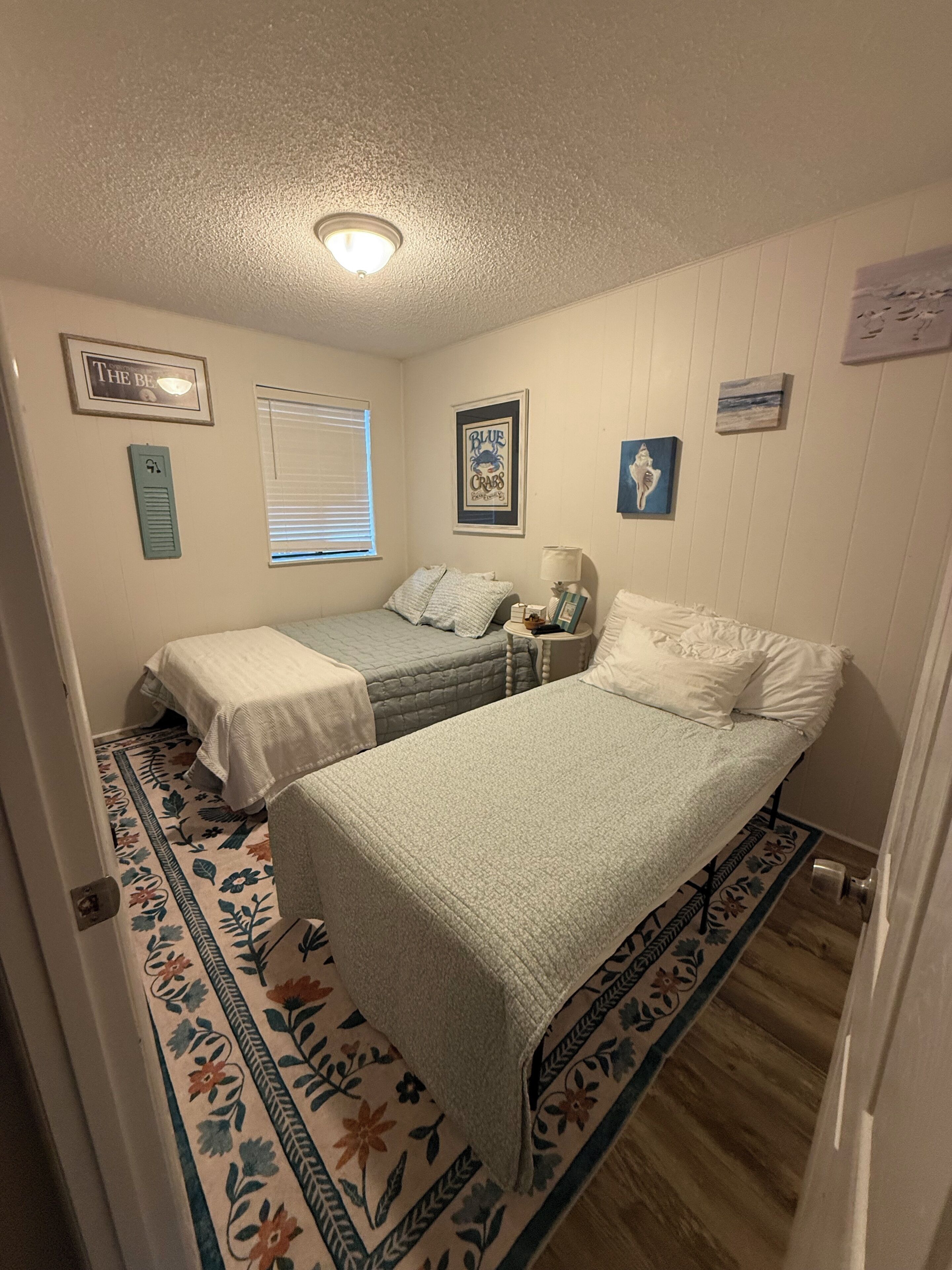 Een slaapkamer met twee bedden, een raam, een plafondlamp en een vloerkleed met bloemenprint.