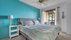 Un dormitorio con pared verde azulado, una cama con colcha estampada, una mesita de noche blanca con lámpara y un ventilador de techo.