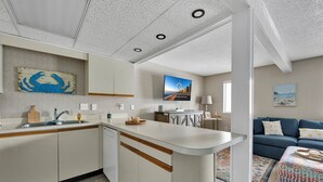 Una cocina moderna con una isla central, un área de comedor con un sofá y una sala de estar con un sofá azul y un reposapiés estampado.