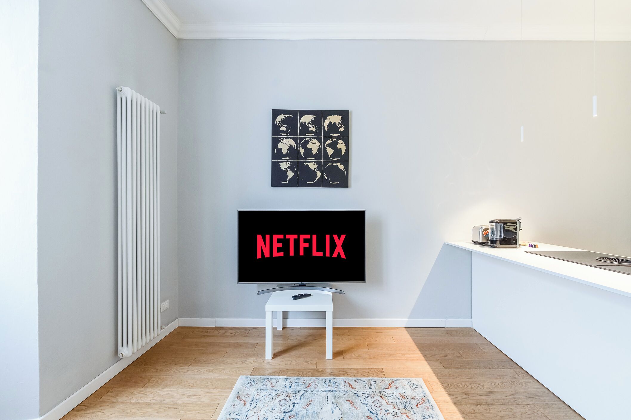 A modern living room with a flat-screen TV mounted on the wall, a white radiator, a small white table, and a kitchen counter with a toaster.