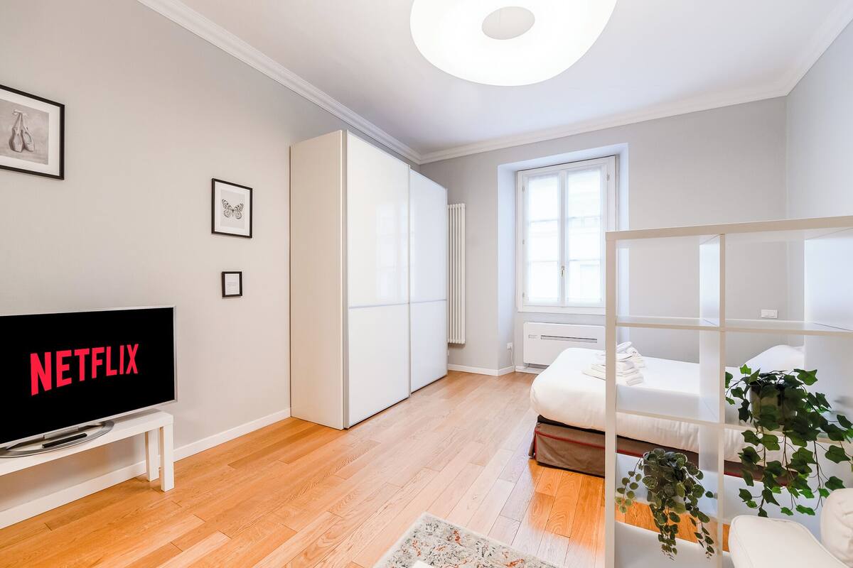 A modern, well-lit room with a flat-screen TV displaying 'NETFLIX', a wardrobe, a bed with a white headboard, and a window with curtains.