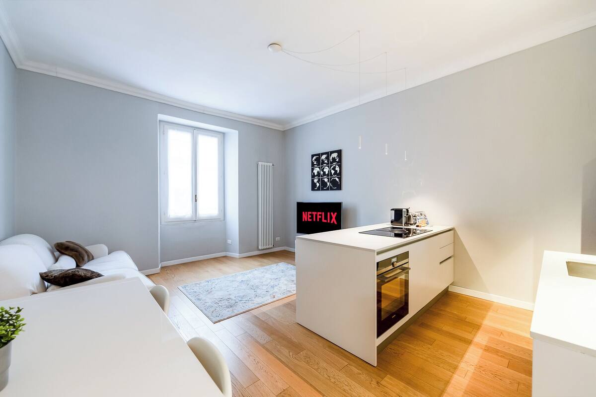 A modern kitchen with a built-in oven, a white countertop, and a small island. There is a window providing natural light and a radiator on the wall.