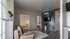 Una sala de estar moderna con un sofá color beige, un televisor de pantalla plana grande montado en una pared oscura y una planta en maceta sobre una estantería.