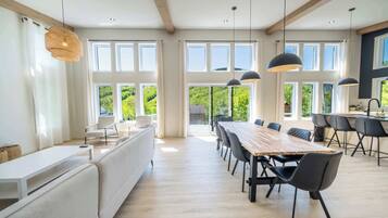 Uma ampla sala de jantar com uma longa mesa de madeira e cadeiras modernas, grandes janelas com vista para o verde, e iluminação pendente.