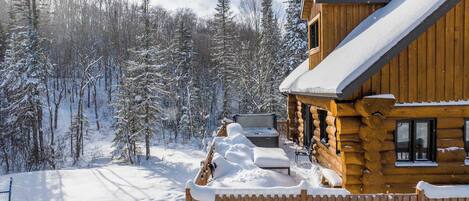 A log cabin with a snow-covered roof, a hot tub, and a wooden fence.
