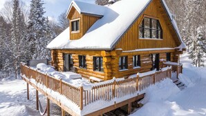 A log cabin with a deck and a snow-covered roof, surrounded by trees. - Spa & Pool table | Le Massif (Saint-Adolphe-d'Howard)