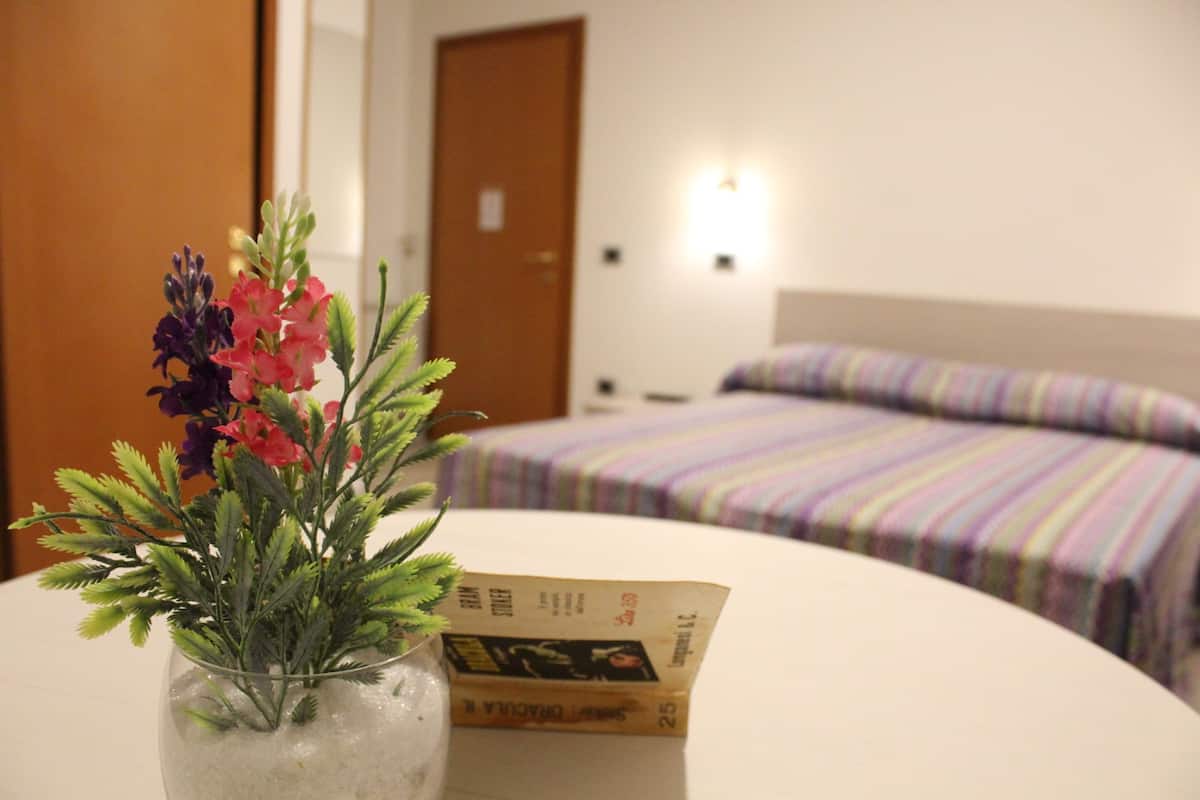 Quarto de hotel com uma cama, mesa de cabeceira com um vaso de flores e uma luminária.