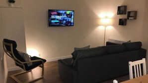 A modern living room with a black sofa, a wooden dining table, and a TV mounted on the wall displaying a program. - 2 Lower Kessock Street (Inverness)