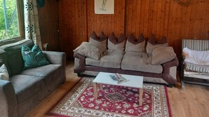 Een gezellige woonkamer met een houten wand, een bank met meerdere kussens, een gestreept fauteuil, een klein tafeltje en een kamerkleed met een patroon.