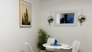 A dining area with a round table, white chairs, a potted plant, and a framed picture of a building.