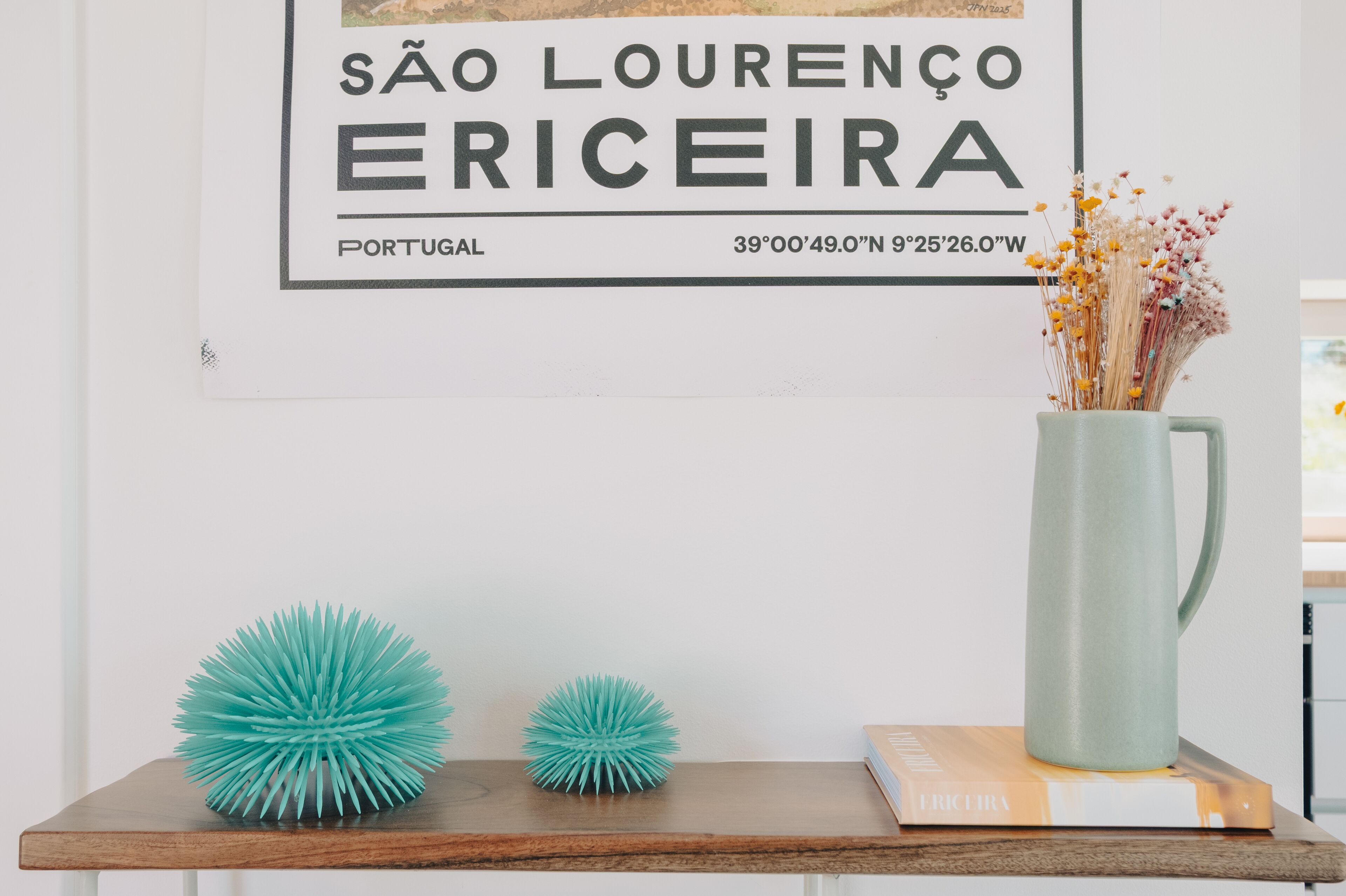 Un cartel enmarco con el texto "SÃO LOURENÇO ERICEIRA, PORTUGAL" y coordenadas, un florero azul turquesa con flores secas, y dos esferas azules sobre una estantería de madera.