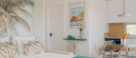 Camera da letto con un letto, una coperta verde a motivi, una parete con un murale di palme, un angolo cottura con un tavolo di legno, sedie e una lampada a sospensione.
