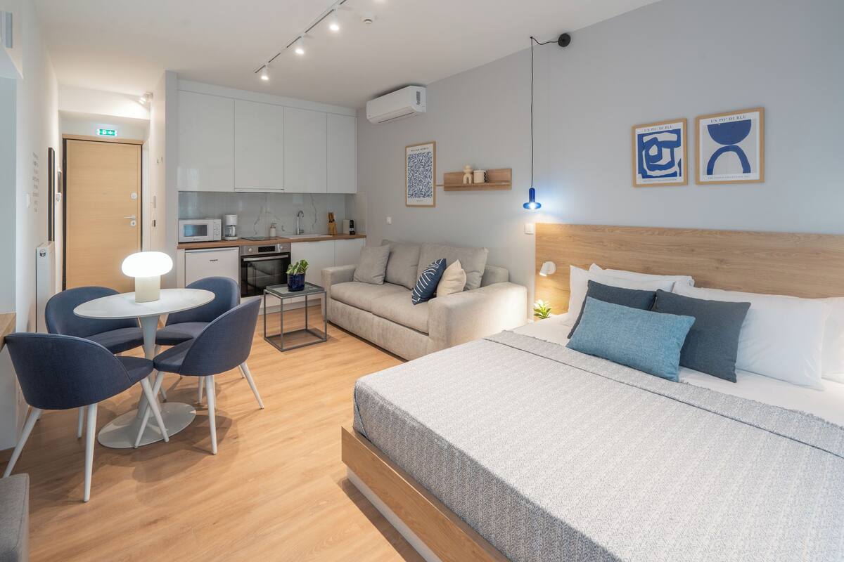 A modern hotel room with a bed, a sofa, a dining table with chairs, a kitchen area, and a small balcony.