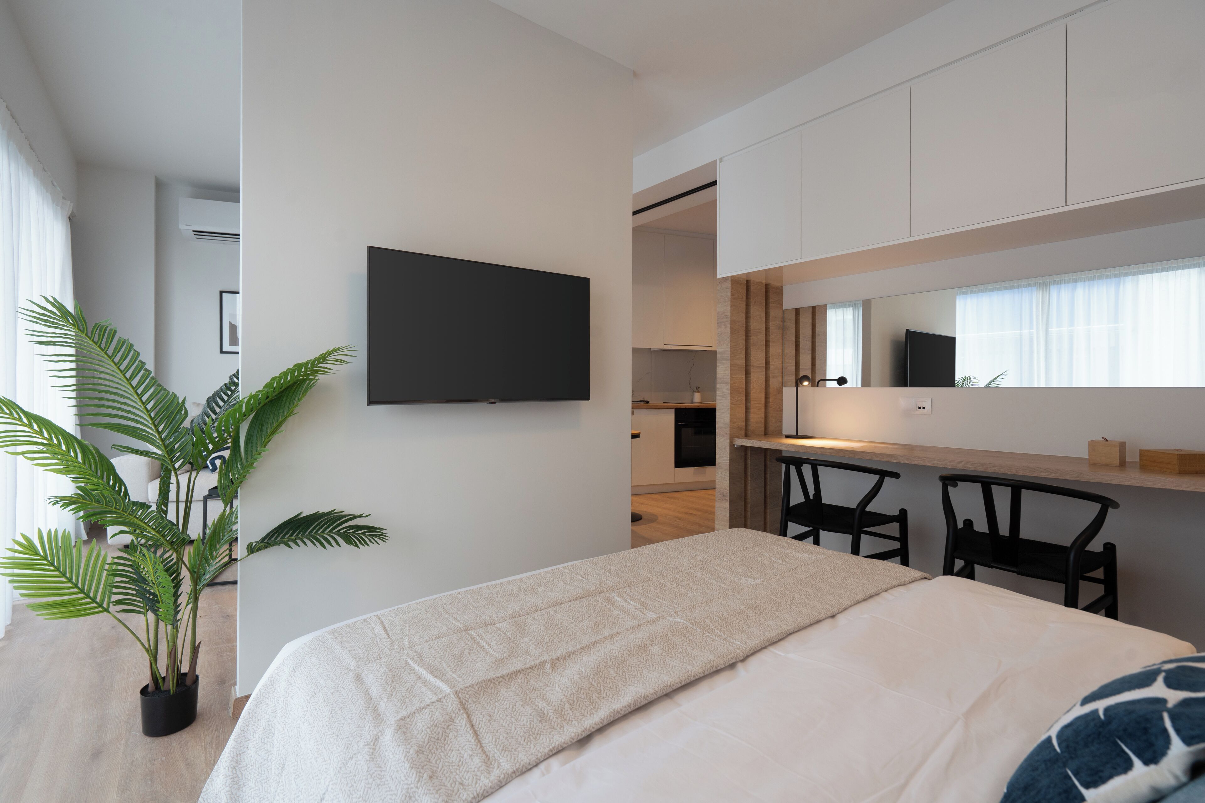 A modern bedroom with a bed, a potted plant, a TV, and a kitchen area in the background.