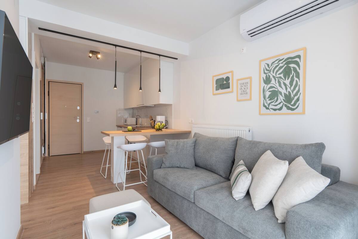 A modern living room with a grey sofa, white coffee table, and a kitchen area in the background.