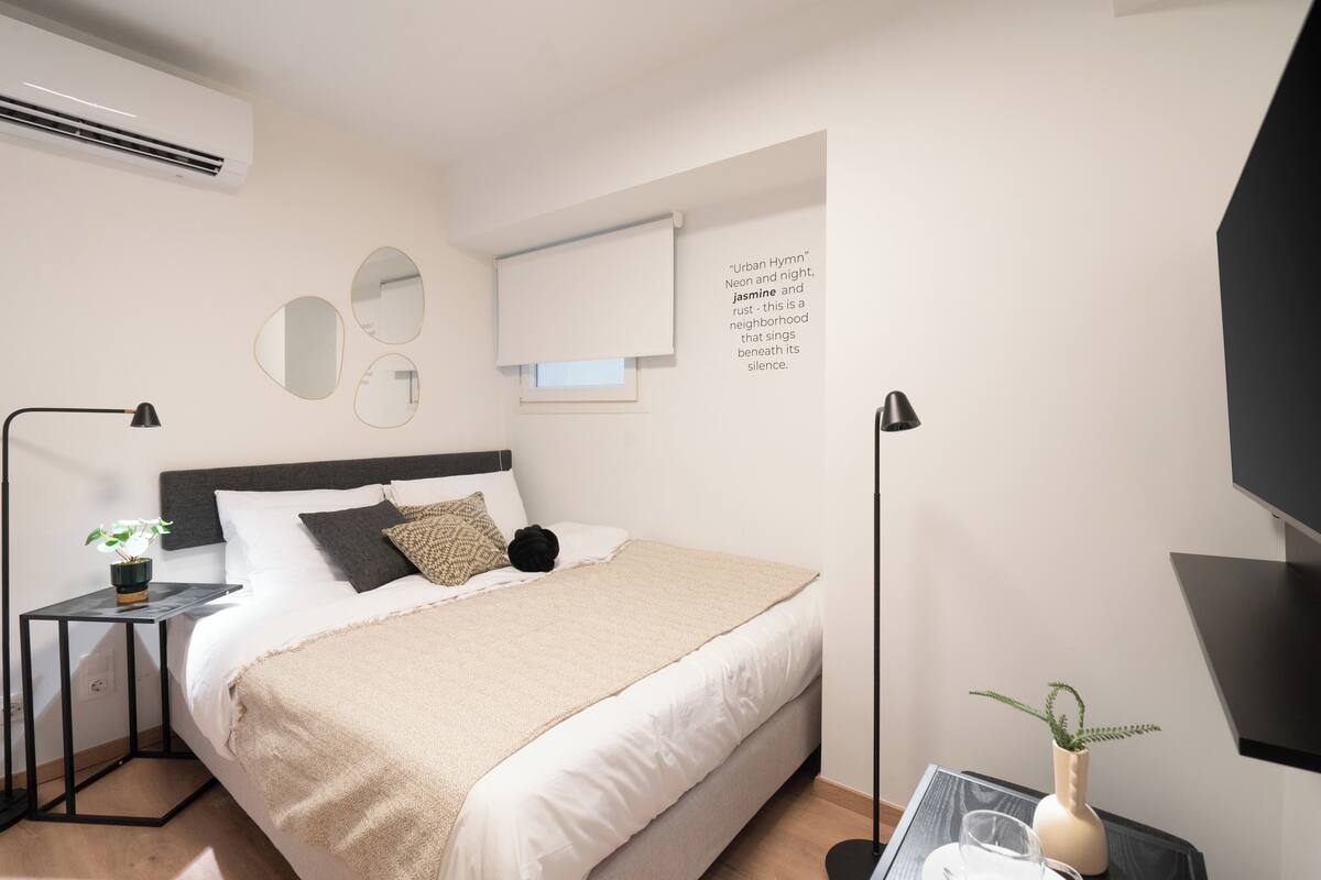A modern bedroom with a bed, bedside tables, a wall-mounted air conditioner, a window with blinds, and a decorative quote.