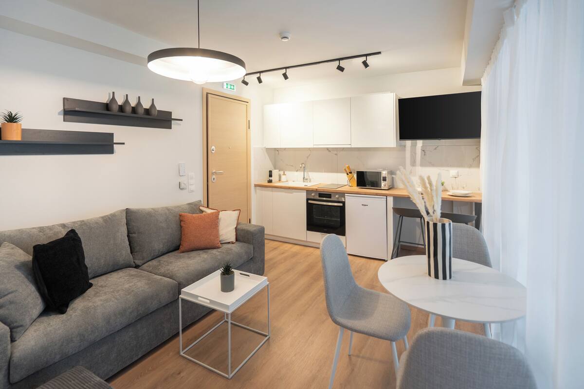 A modern living room with a grey sofa, a small white table, and a grey chair. There is a kitchen area with white cabinets, a microwave, and a sink.