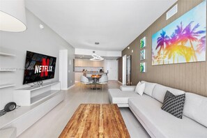 A modern living room with a flat-screen TV, a wooden coffee table, a sofa with patterned cushions, and a dining area with a white table and chairs.