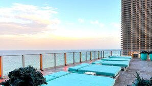 Una terrazza panoramica con diversi lettini azzurri e piante in vaso, con vista sull'oceano e un grattacielo.