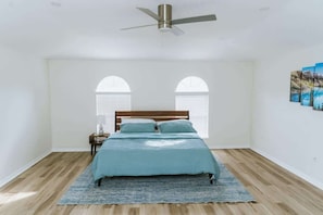 A bedroom with a bed, a ceiling fan, two windows with arched tops, and a painting on the wall. - Beachside Bliss: 4BR Home in Palm Coast Paradise (Palm Coast)