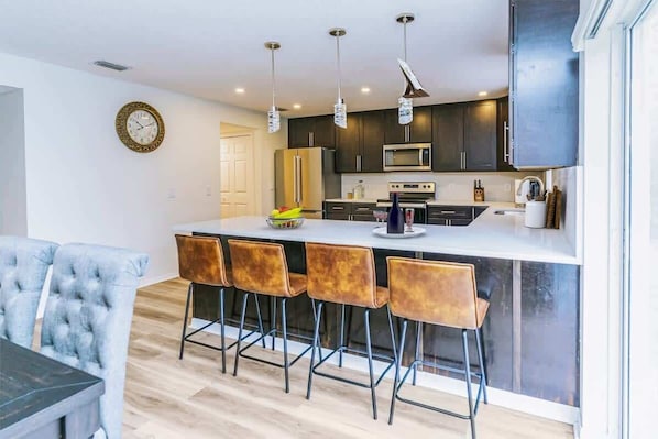 A modern kitchen with dark cabinets, a central island with bar stools, a microwave, and a clock on the wall. - Beachside Bliss: 4BR Home in Palm Coast Paradise (Palm Coast)