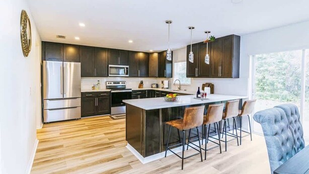 A modern kitchen with dark cabinets, stainless steel appliances, a central island with bar stools, and a large window providing natural light. - Beachside Bliss: 4BR Home in Palm Coast Paradise (Palm Coast)