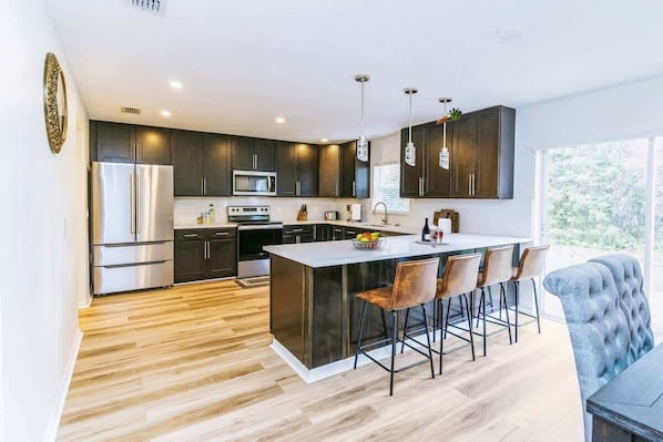A modern kitchen with dark cabinets, stainless steel appliances, a central island with bar stools, and a large window providing natural light.