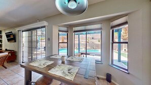 Ein geräumiges Wohnzimmer mit einem großen Holztisch, einem an der Wand montierten Fernseher und großen Fenstern mit Blick auf einen Hinterhof und einen Pool.