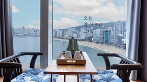 A room with a large window overlooking a cityscape and beach. There is a wooden table with a tray containing two cups and a green object. Two chairs are positioned in front of the table.