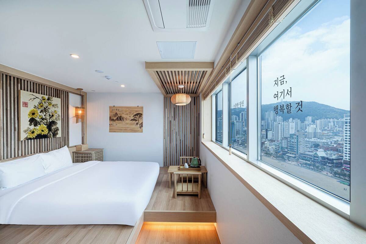 Una habitación de hotel moderna con una cama grande, una mesita con un hervidor, vista a la ciudad y un cuadro en la pared.