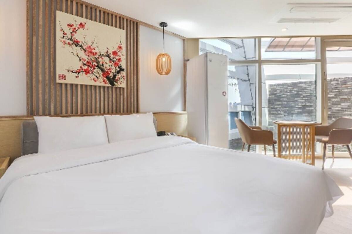 Habitación de hotel con una cama grande, un cuadro en la pared, una mesa con sillas y vistas de la ciudad a través de amplias ventanas.
