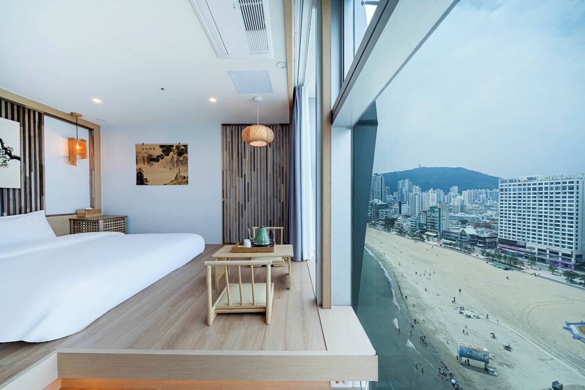 Habitación de hotel moderna con una cama grande, una mesita de madera con un hervidor y vistas a la playa y al paisaje urbano.