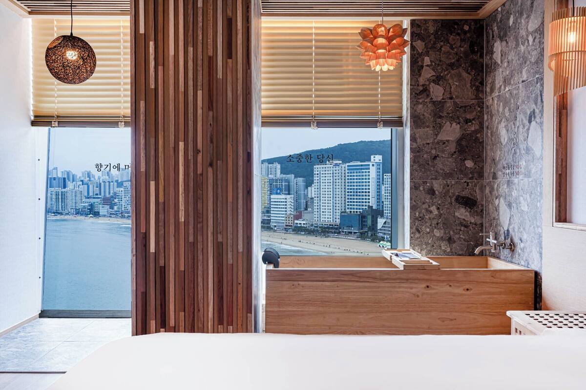 Un baño moderno con tocador de madera, pared de acento de piedra y vistas a un paisaje urbano a través de amplias ventanas.