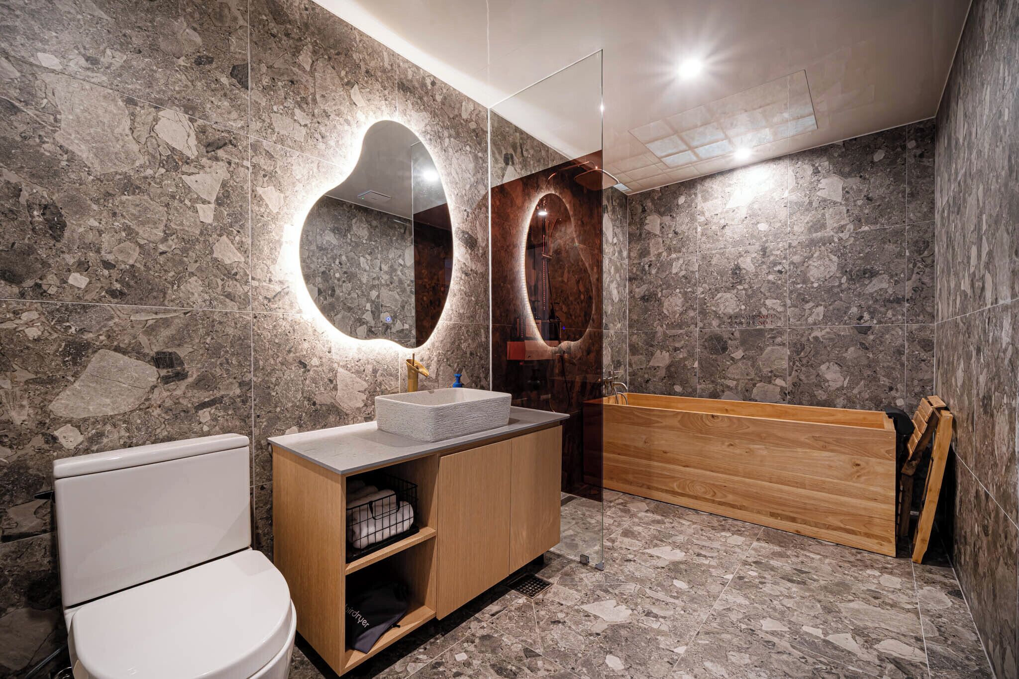 Un baño con pared de mármol, tocador de madera, inodoro blanco y bañera de madera.