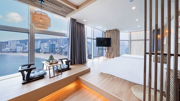 A modern hotel room with a large window offering a city view, a flat-screen TV, a sofa, and a small table with decorative items.