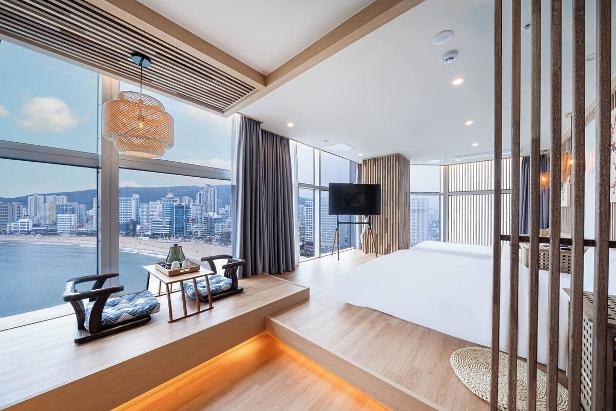 Una habitación de hotel moderna con un ventanal que ofrece vistas a la ciudad, un televisor de pantalla plana, un sofá y una mesita con objetos decorativos.