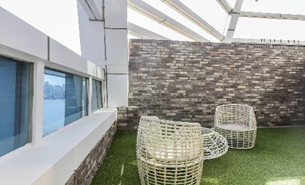 A modern outdoor area with a glass wall, artificial grass, wicker furniture, and a brick wall.