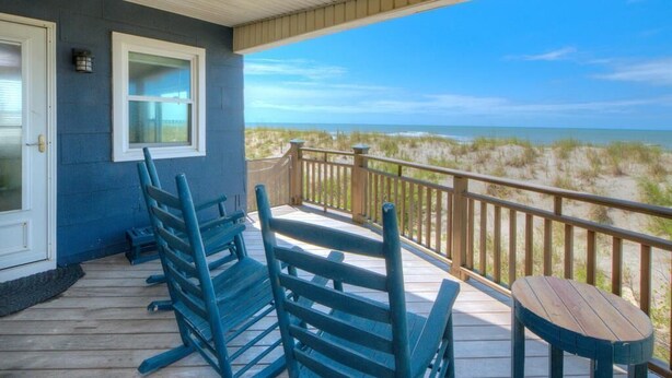 Eine Holzterrasse mit blauen Stühlen und einem Tisch, mit Blick auf einen Strand mit Dünen.