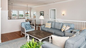 Un vaste salon avec un canapé gris, une table basse en bois et un fauteuil en osier agrémenté d’un coussin bleu. On y trouve également un coin repas avec une table et des chaises, une lampe, et une vue sur l’extérieur grâce à de grandes fenêtres.